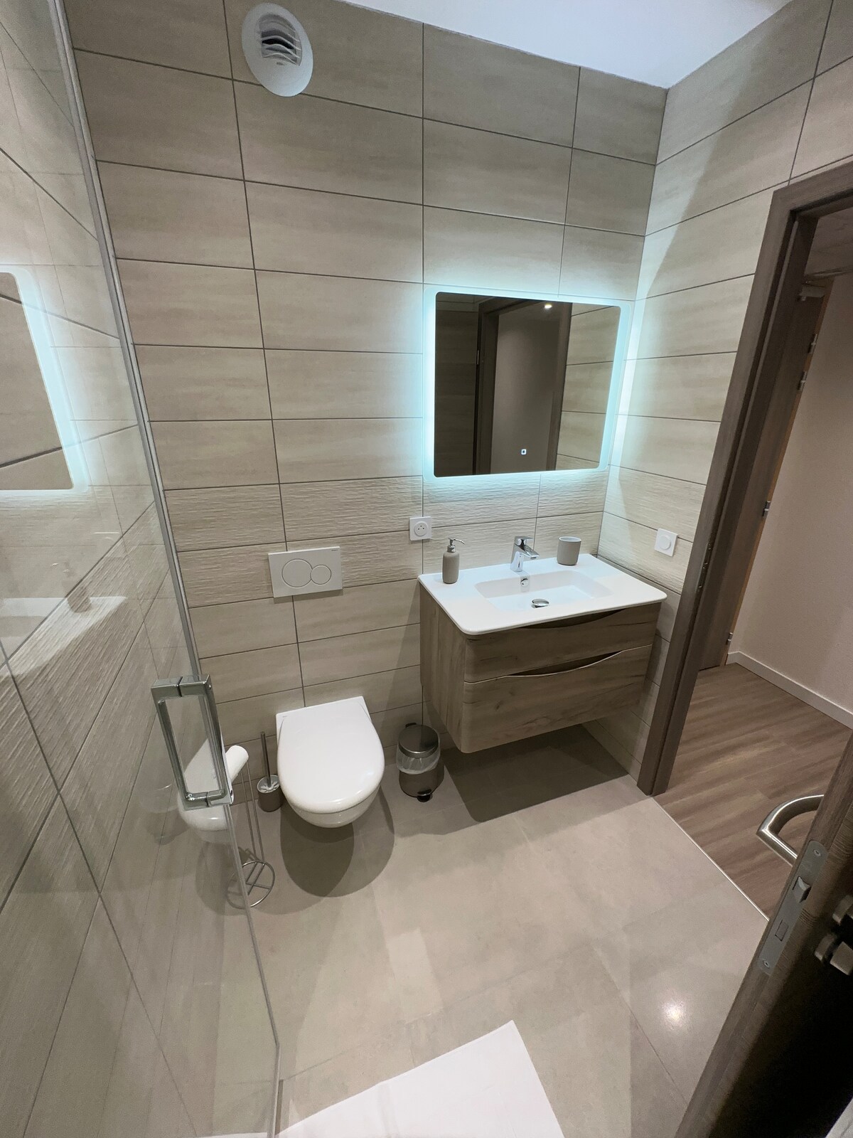 A modern bathroom features wood accents and neutral tiles, providing a clean and sleek look. A compact vanity is placed under an illuminated mirror, with toiletries strategically displayed. The shower area is enclosed by glass, while a white toilet is positioned alongside.