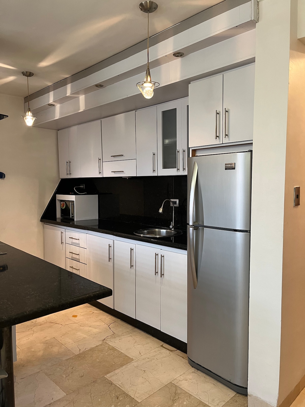 The kitchen area features modern cabinetry with sleek white fronts, complemented by dark granite countertops. Stainless steel appliances, including a refrigerator and microwave, are present. Pendant lighting hangs from the ceiling, adding subtle illumination to the space.