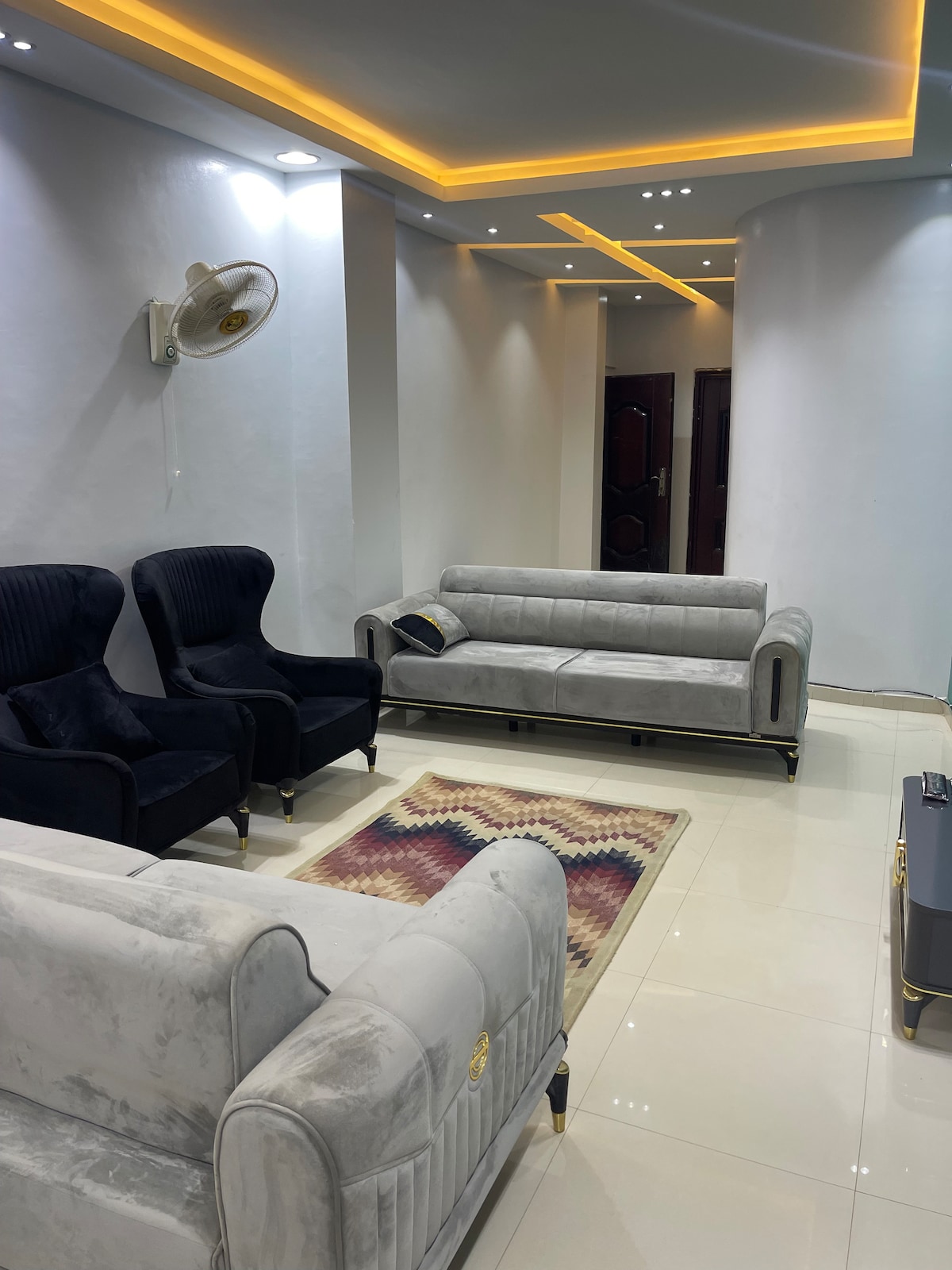 A modern living area features a light-colored sofa and two stylish black armchairs. A decorative area rug lies on the polished tile floor, while soft ceiling lights provide illumination. The entrance to another room is visible in the background.