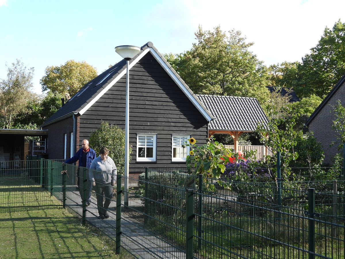 A charming dark wooden house is surrounded by a well-maintained garden, featuring various plants and trees. A pathway leads to the entrance, where a couple is seen walking together. Sunlight filters through the leaves, creating a peaceful outdoor setting.