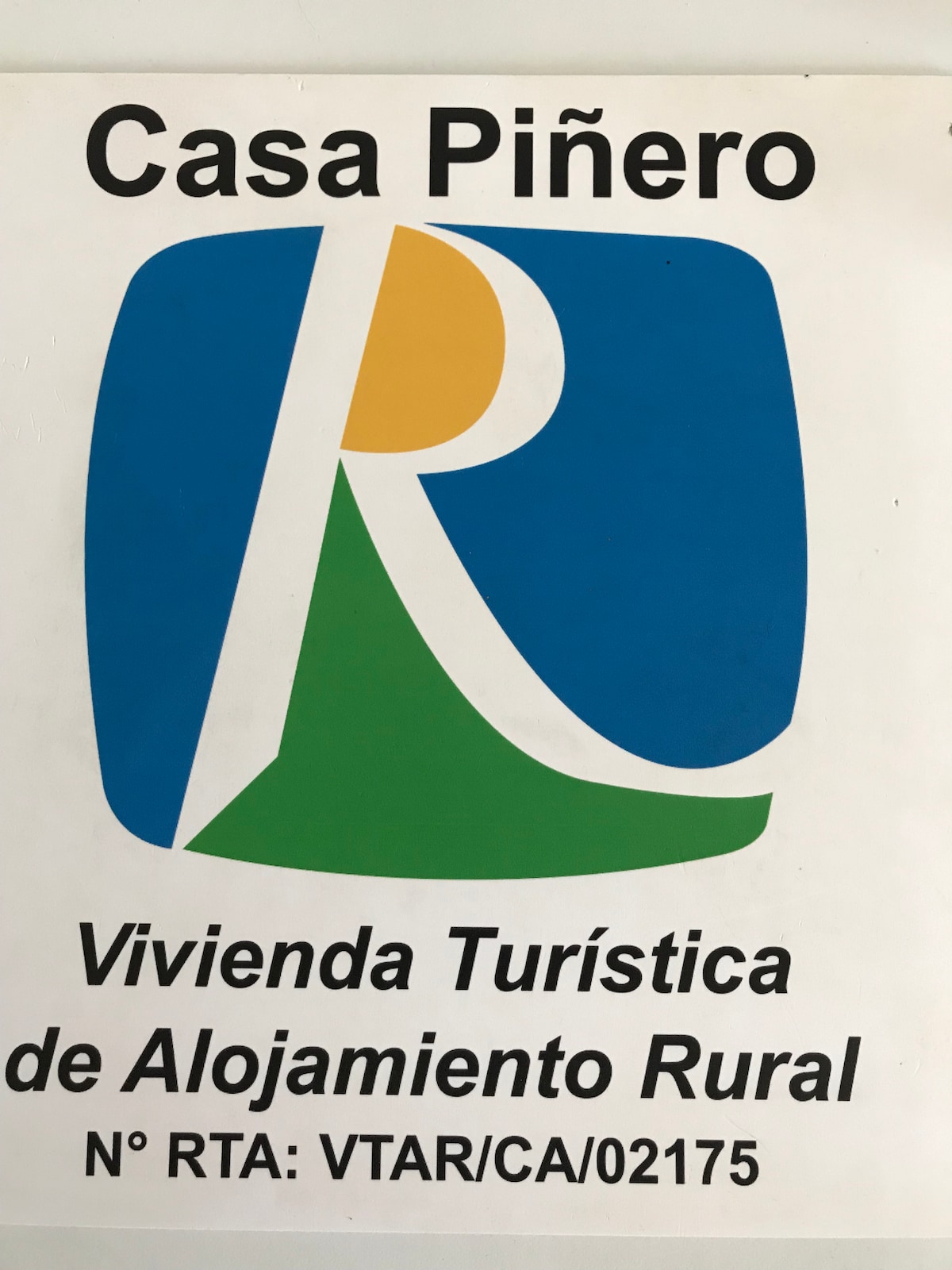A sign for Casa Piñero is displayed prominently, featuring a colorful logo with a stylized letter 'R' alongside vibrant shapes in blue, green, and yellow. The text indicates it as a rural tourist accommodation, noted with a registration number for authenticity.