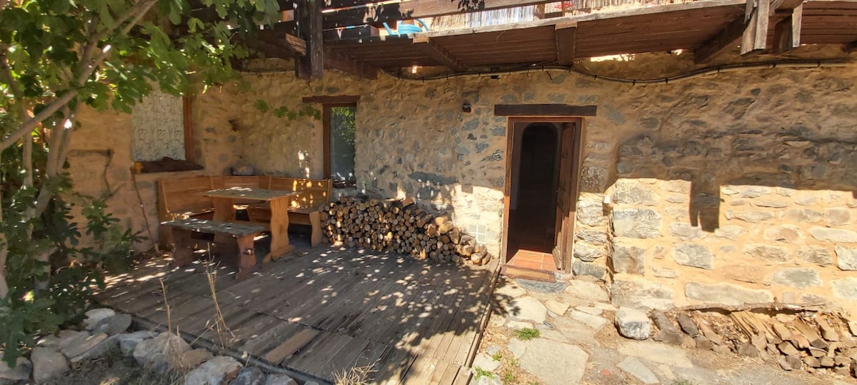 An outdoor seating area features a wooden table and benches, positioned alongside a stack of firewood. The rustic stone walls offer a natural backdrop, while the wooden deck is lined with stones, creating a cozy and inviting space.