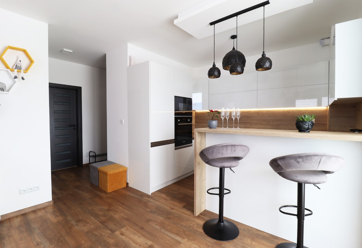 A modern kitchen is highlighted by a sleek countertop with two stylish bar stools. Above, three pendant lights provide illumination. A built-in oven and cabinetry are visible, complemented by wooden elements, creating a clean and inviting space.