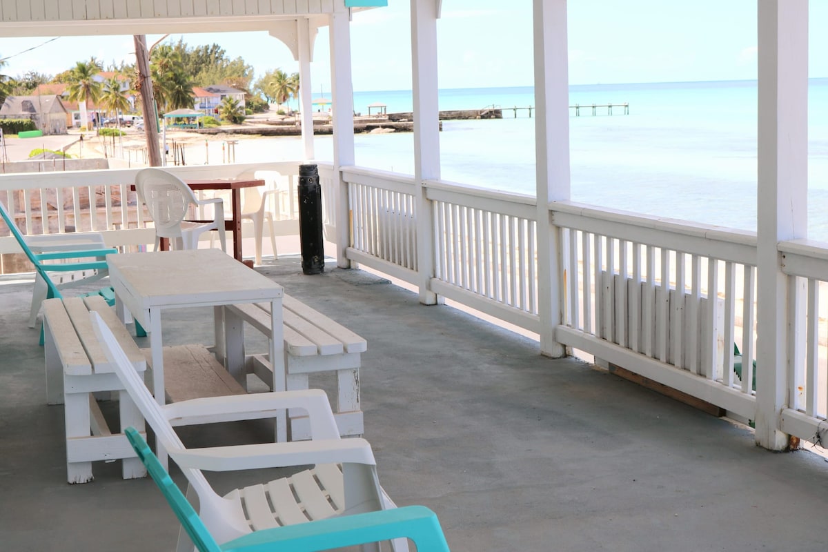 A spacious porch area with wooden flooring overlooks the beach. Several white and turquoise chairs are arranged near a wooden table. The railing provides an unobstructed view of calm waters and distant palm trees, creating a serene outdoor setting.