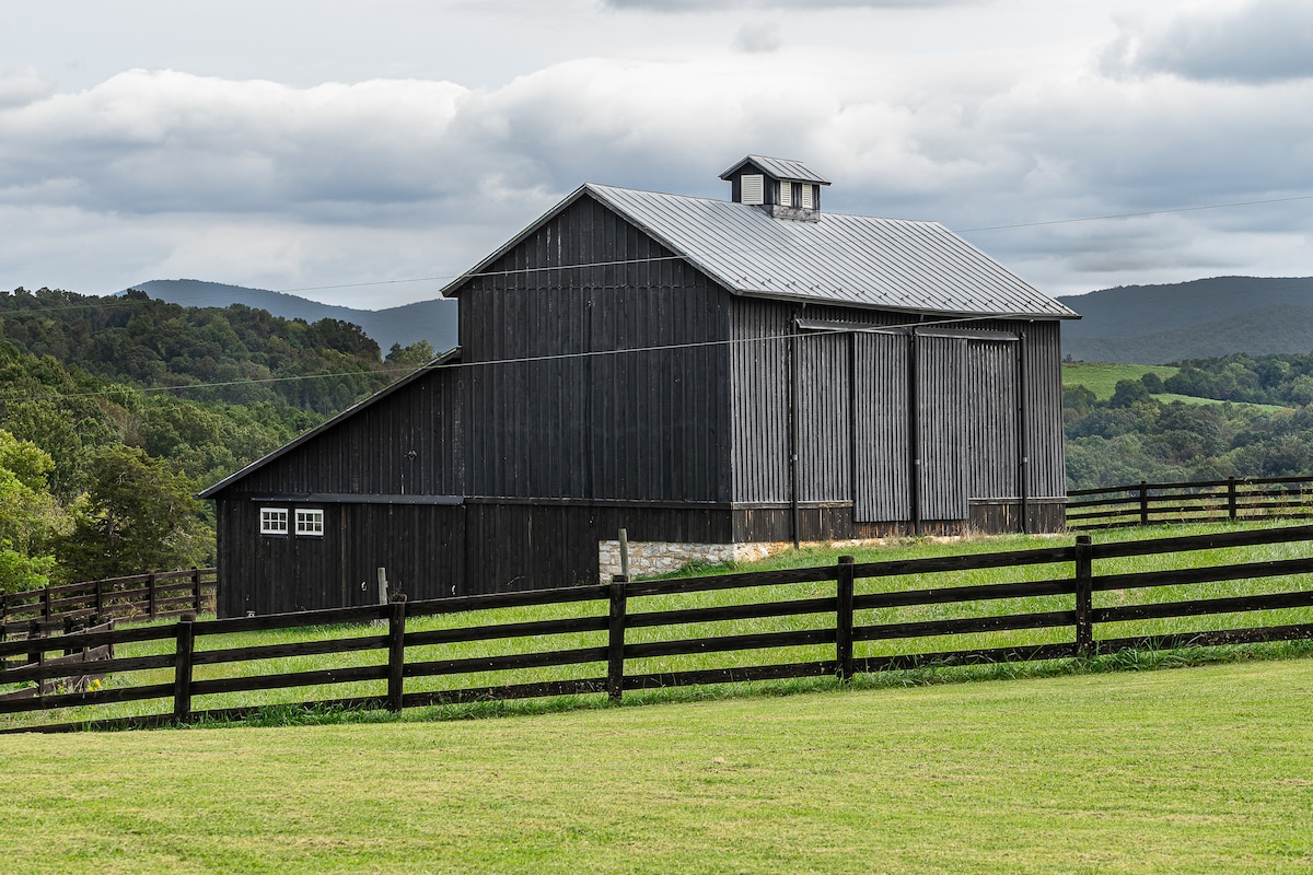 A large, black barn with a metal roof is set against a backdrop of rolling green hills. Wooden fencing outlines the property, and the cloudy sky creates a serene atmosphere. Windows and sliding doors are visible on the barn's facade, highlighting its rustic charm.