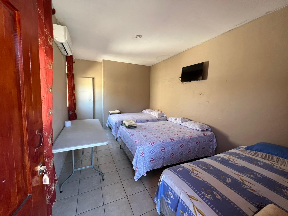 The room features three beds arranged neatly, each with light bedding. A small table is positioned beside the beds, and a wall-mounted TV is present. Natural light enters through a window, while a door leads to the adjacent area.