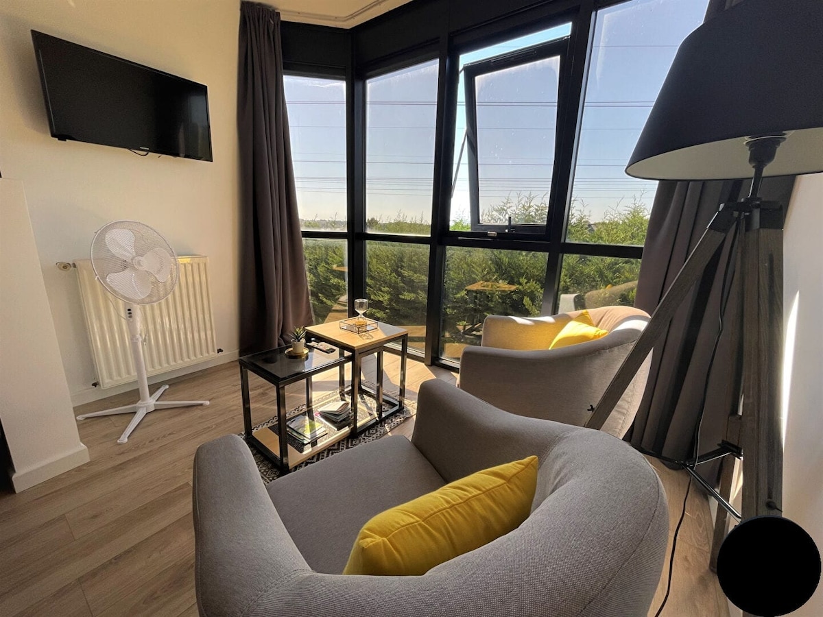 A cozy seating area features two armchairs with yellow cushions and a small table. A flat-screen TV is mounted on the wall, while a floor lamp adds soft illumination. Large windows offer a view of greenery, allowing natural light to brighten the space.