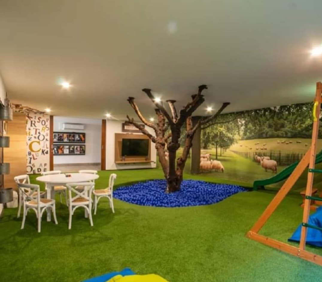 A playful children's playroom is featured, featuring a decorative tree in the center and a carpet of green turf. Soft seating areas surround a circular table, while a slide and climbing structure are present. A mural depicting sheep in a pastoral scene complements the space.