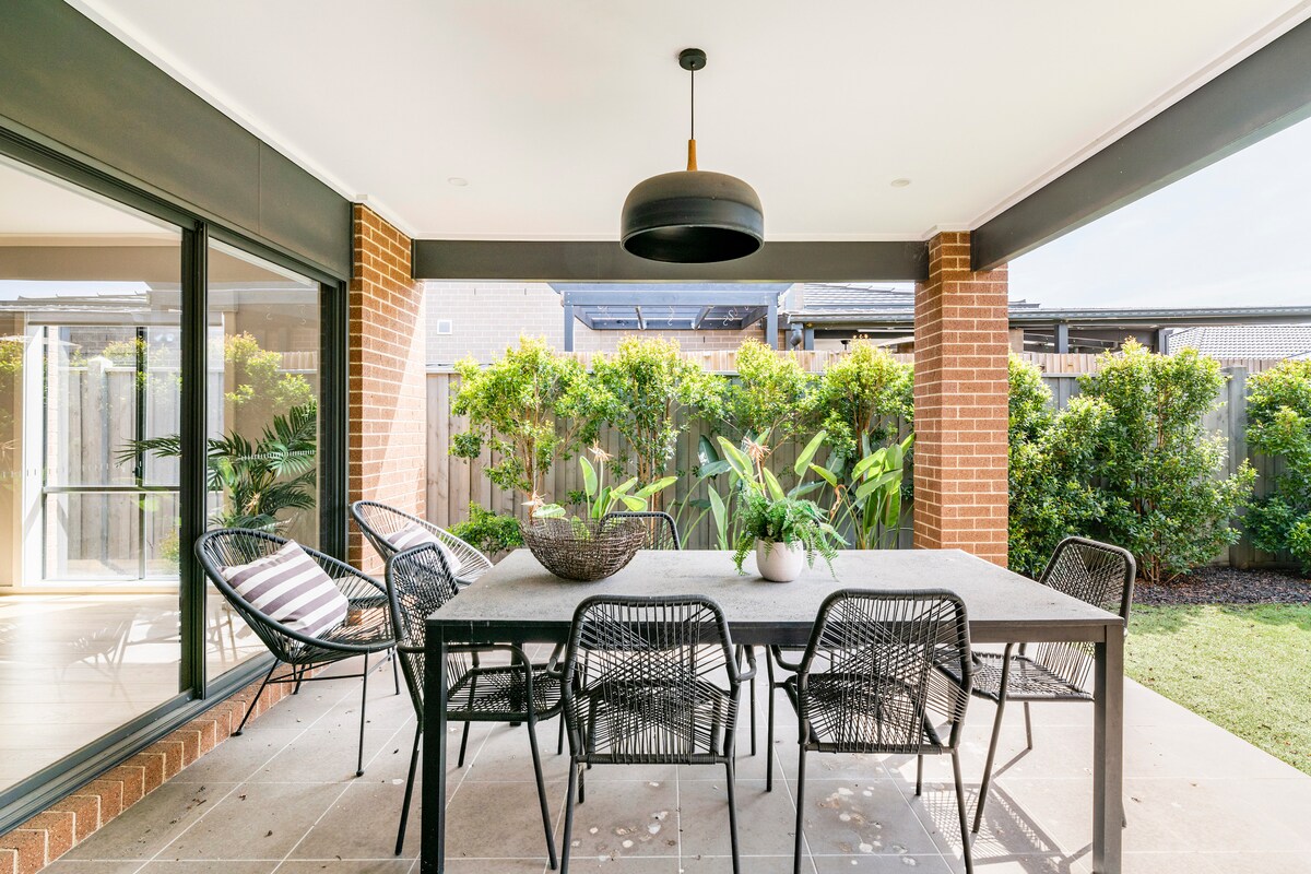 An outdoor dining area is observed, featuring a rectangular table surrounded by six woven chairs. A hanging light fixture is suspended above the table. Lush greenery is visible in the background, with a patio area constructed from light-colored stone.