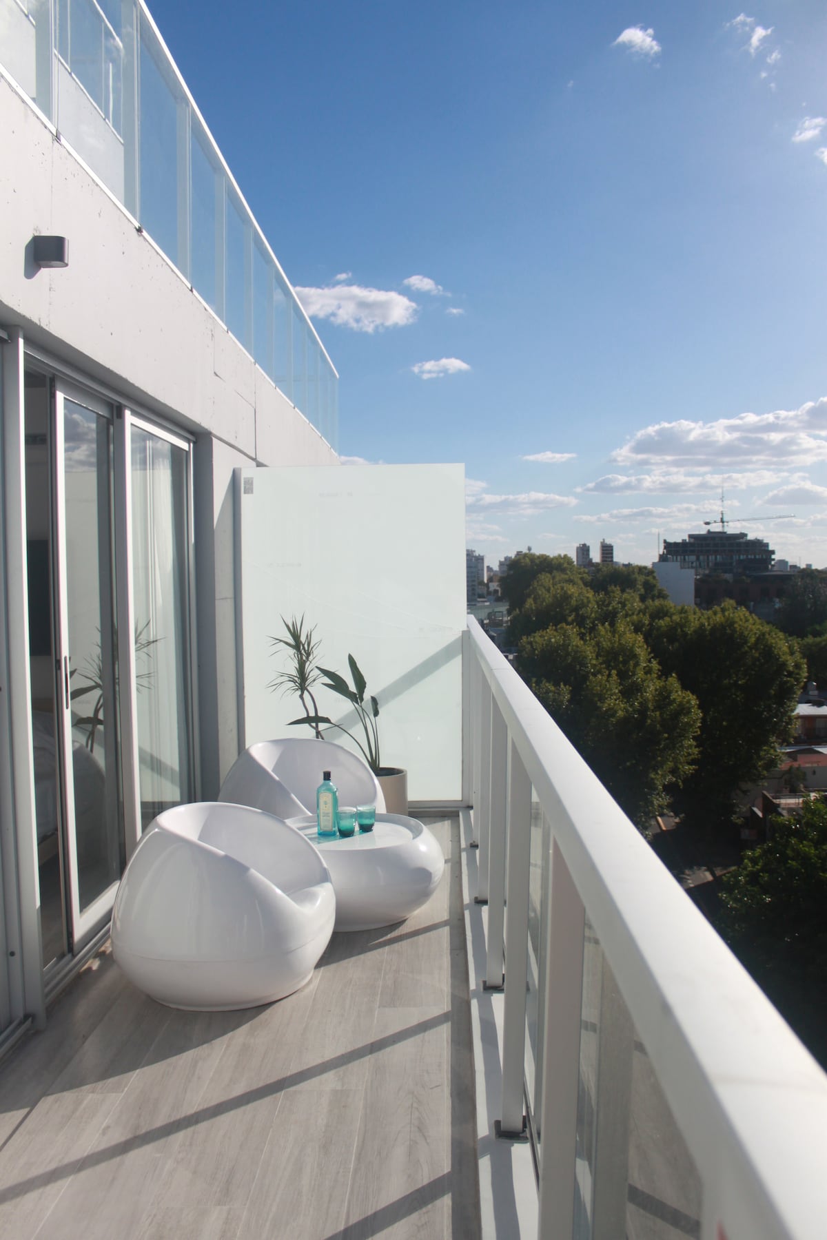 A spacious balcony is shown, featuring modern white chairs and a small potted plant. Large glass doors lead into the interior space, while the view offers a glimpse of greenery and cityscape under a clear blue sky.