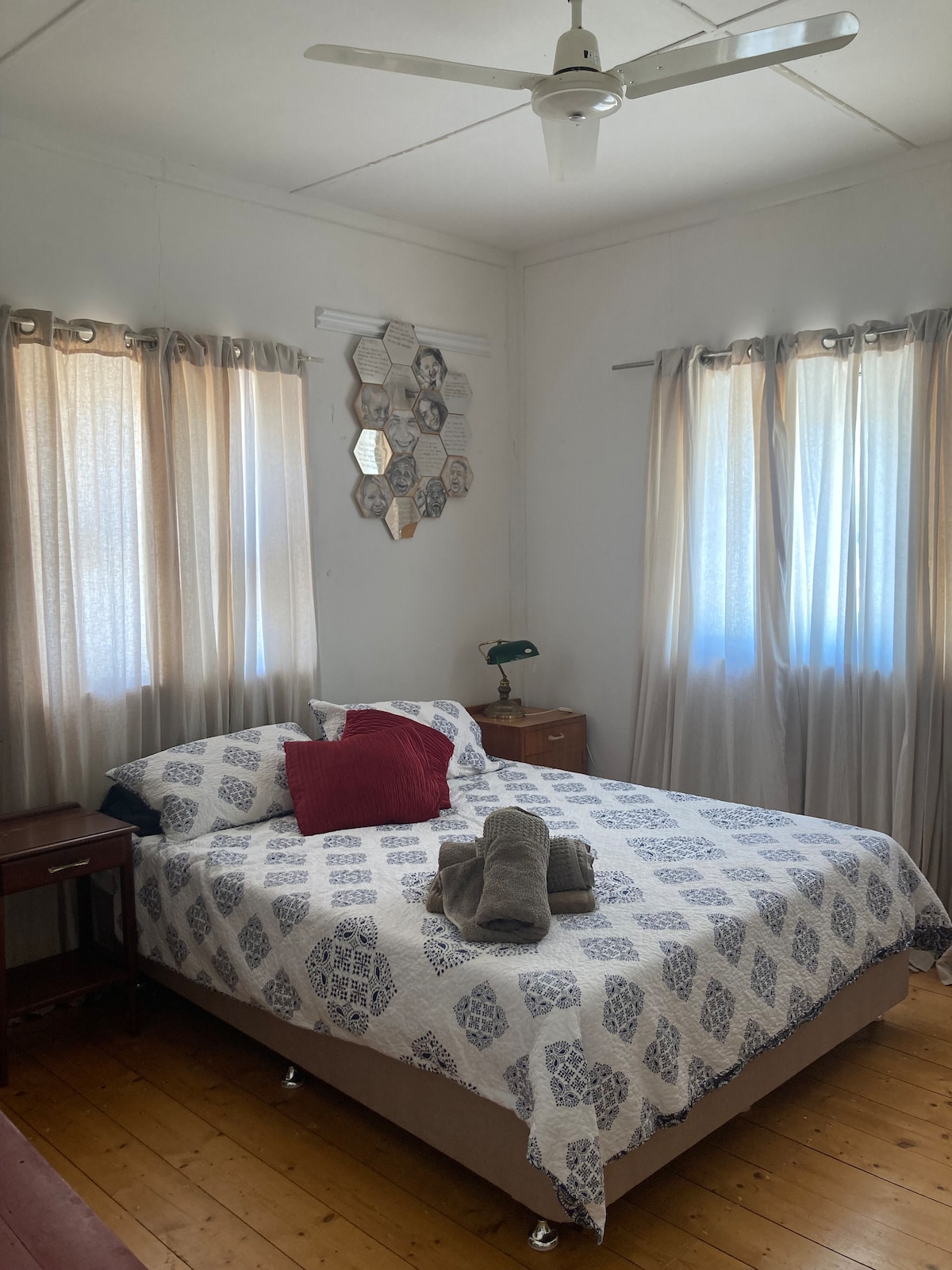 A well-lit bedroom features a spacious bed decorated with a patterned bedspread. Soft, neutral-toned curtains frame two windows, allowing natural light to enter. A bedside table holds a lamp, and neatly rolled towels are placed on the bed for convenience.