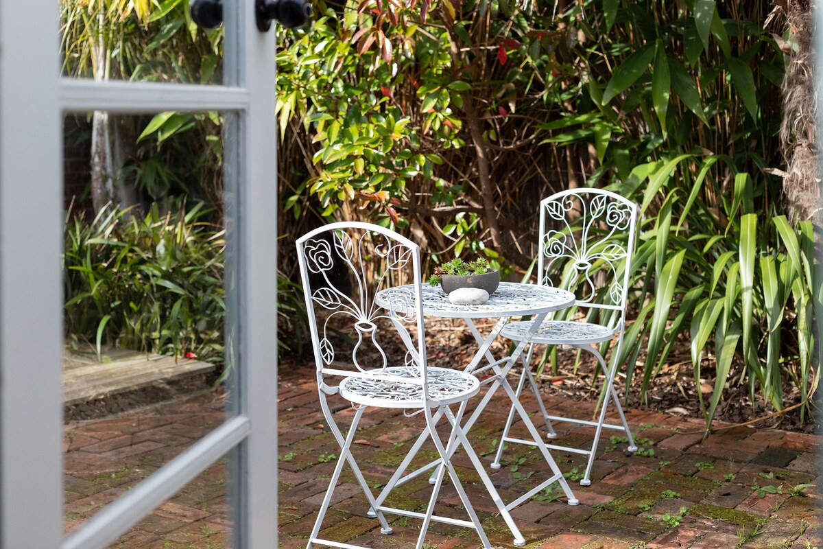 A private courtyard is depicted, featuring a small bistro set with a round table and two folding chairs made of white metal. Lush greenery surrounds the space, providing a tranquil atmosphere. The setting invites relaxation and enjoyment of a morning coffee.