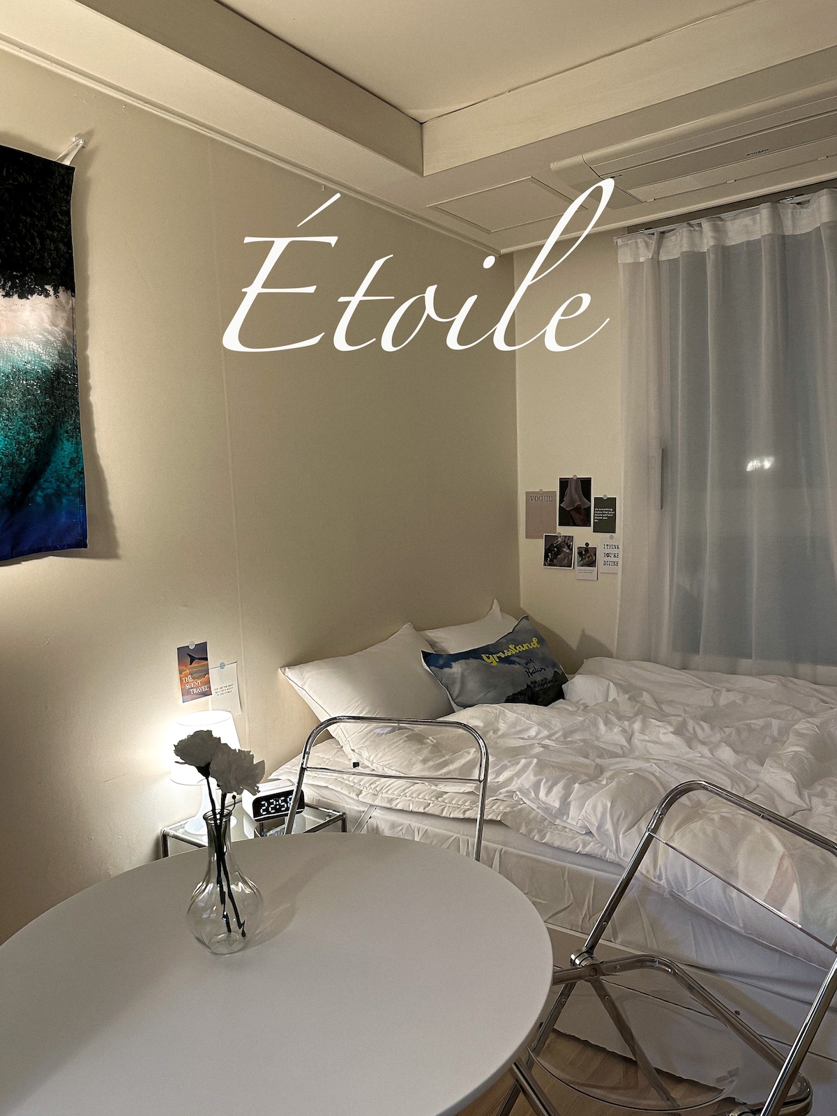 A cozy bedroom features a queen-size bed with clean, white linens. A small table with a glass vase and flowers is positioned nearby. Natural light filters through sheer curtains, complementing a wall adorned with photographs and art.