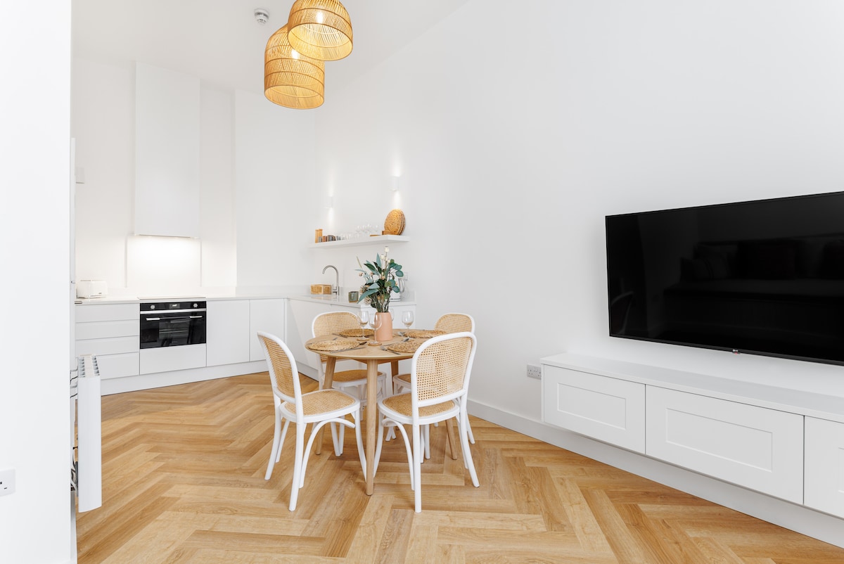 A light-filled dining area features a round wooden table surrounded by four white chairs. The kitchen is equipped with sleek, modern appliances. Wood flooring in a herringbone pattern leads the eye towards a spacious lounge area, creating an inviting ambiance.