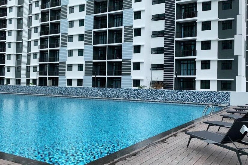 An expansive swimming pool is framed by a surrounding deck, with several lounge chairs positioned for relaxation. The building's modern architecture features multiple floors and windows, providing a contemporary backdrop to the serene blue water.