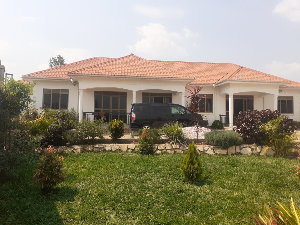 A spacious single-story building with a red-tiled roof is surrounded by well-maintained gardens. Lush green lawns and a variety of colorful plants frame the entrance, while large windows offer ample natural light. A parked vehicle is visible in the driveway, enhancing accessibility.