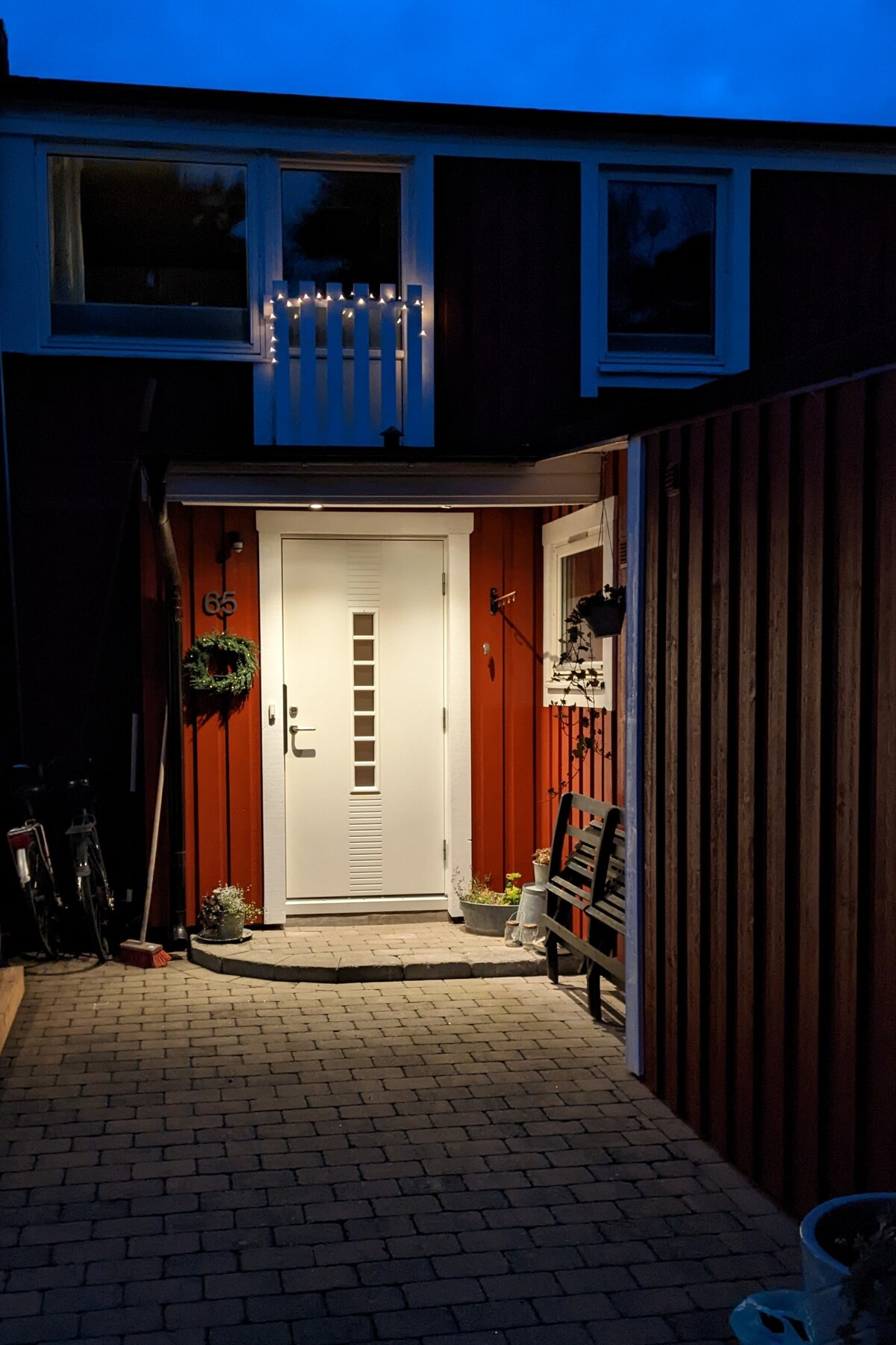 The entrance of the house is softly illuminated, featuring a modern front door with decorative glass panels. A welcoming atmosphere is created by the subtle lighting on the exterior, while a small bench and potted plants add to the inviting entryway.