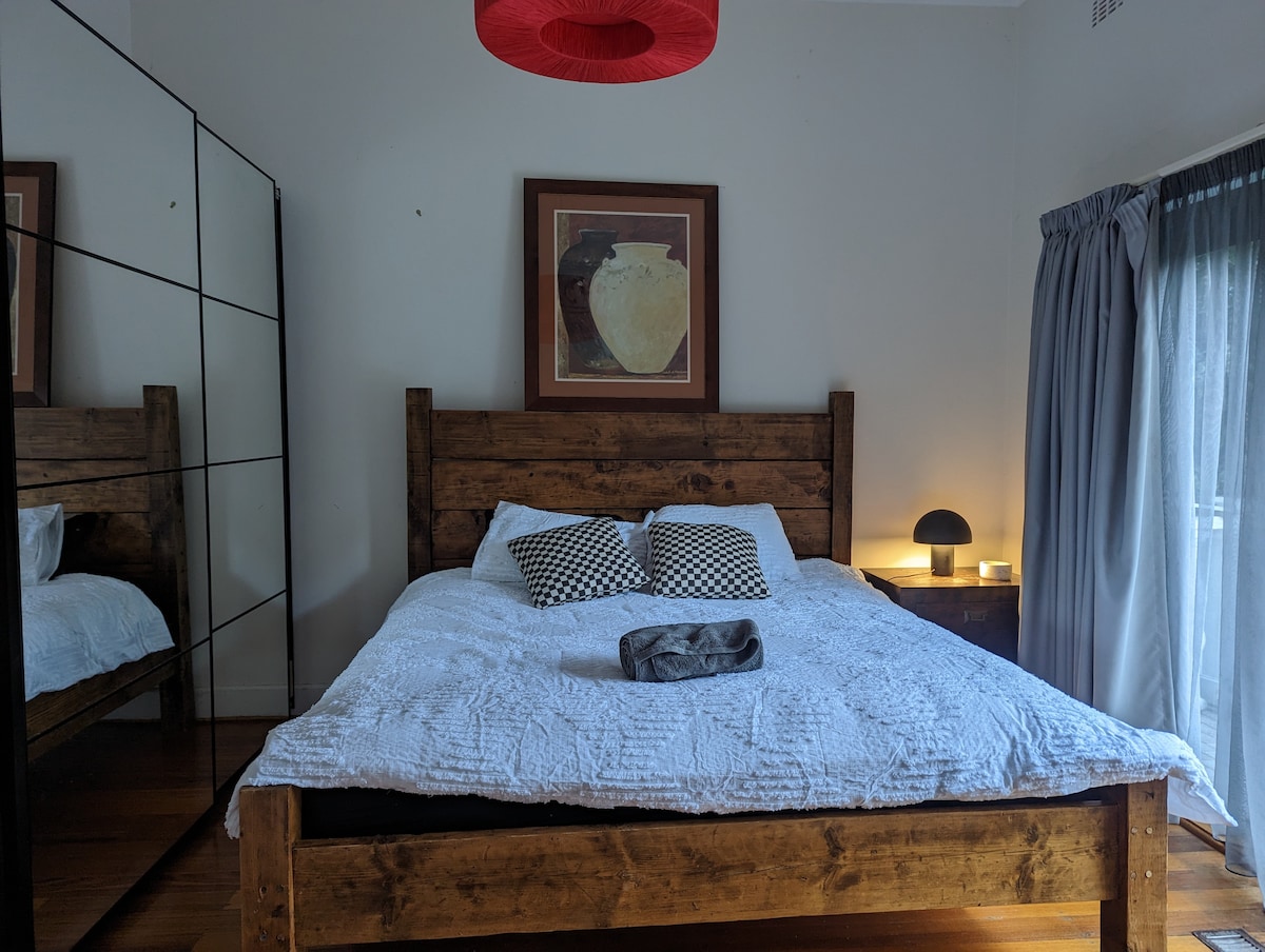 A spacious sleeping area features a wooden bed frame with a cozy quilt and two patterned pillows. A large mirrored wardrobe reflects light, and a soft lamp sits on a nearby nightstand. Sheer curtains allow natural light to brighten the neutral-colored room.