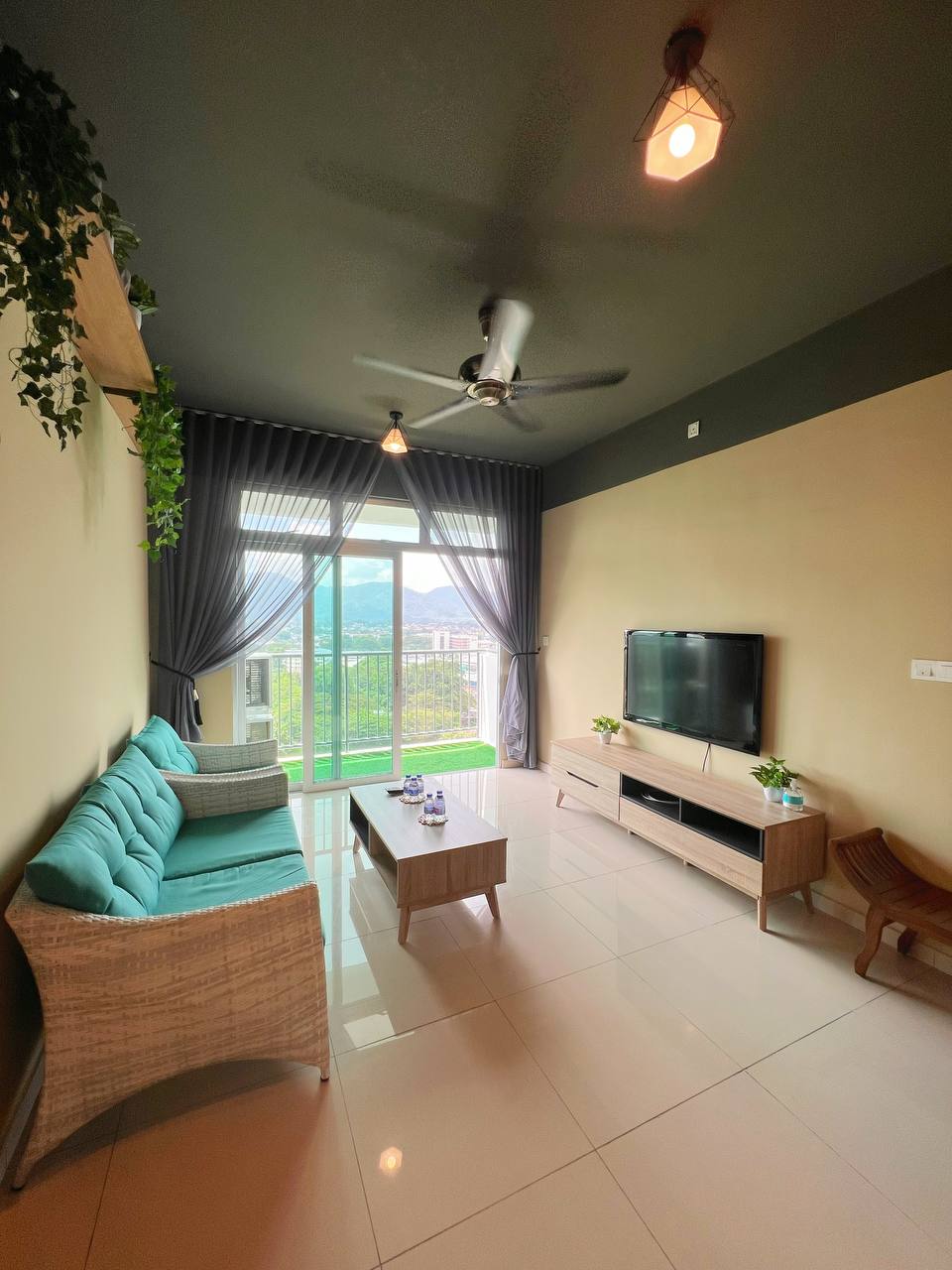 A well-lit living space features a comfortable seating area with a turquoise couch and a minimalist coffee table. Large glass doors allow natural light to fill the room, revealing views of the outside. A television is mounted on the wall opposite the seating, enhancing the indoor experience.