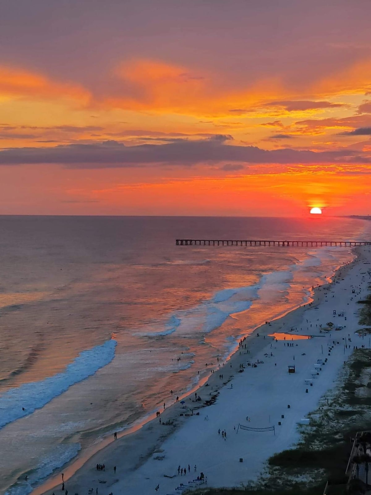A breathtaking view of the sunset over the ocean is captured, with sun rays reflecting on the water. A sandy beach extends along the shore, where small figures can be seen enjoying the shoreline. A pier stretches out into the sea, enhancing the picturesque coastal scenery.