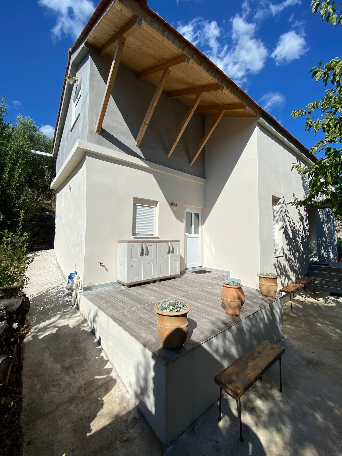 An exterior view of the country villa showcases a light-colored façade and a wooden overhang. The deck area features potted plants and a wooden bench. Clear blue skies and scattered clouds create a bright atmosphere, highlighting the surrounding olive grove.