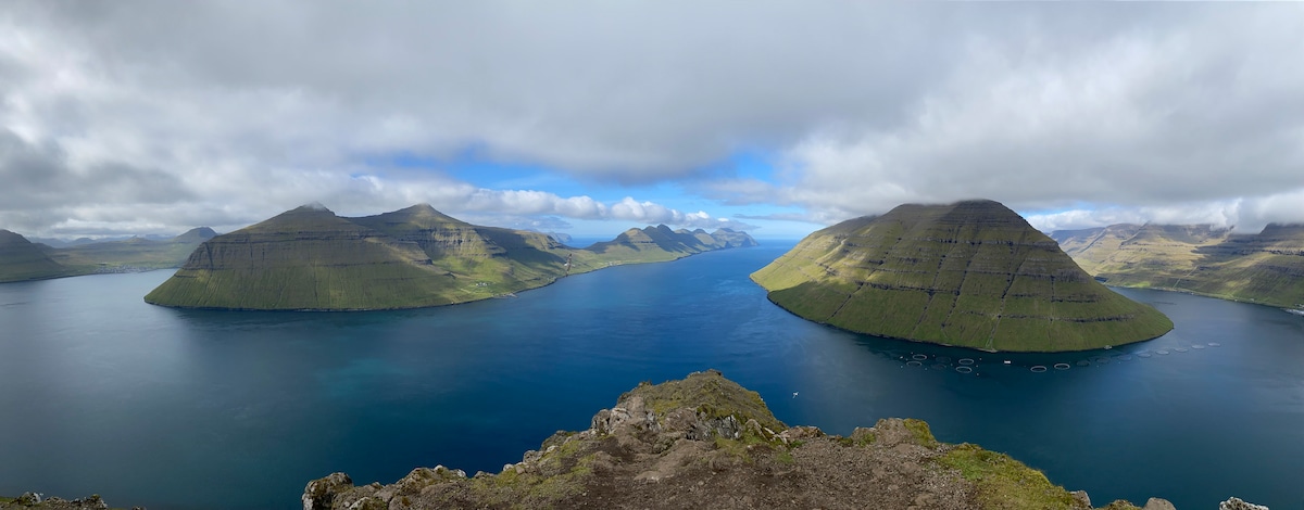A panoramic view of the surrounding islands is captured, showcasing steep green hills rising from tranquil waters. The scene is framed by a partly cloudy sky, allowing glimpses of blue. The landscape displays varying shades of vibrant green, characteristic of the area's natural beauty.