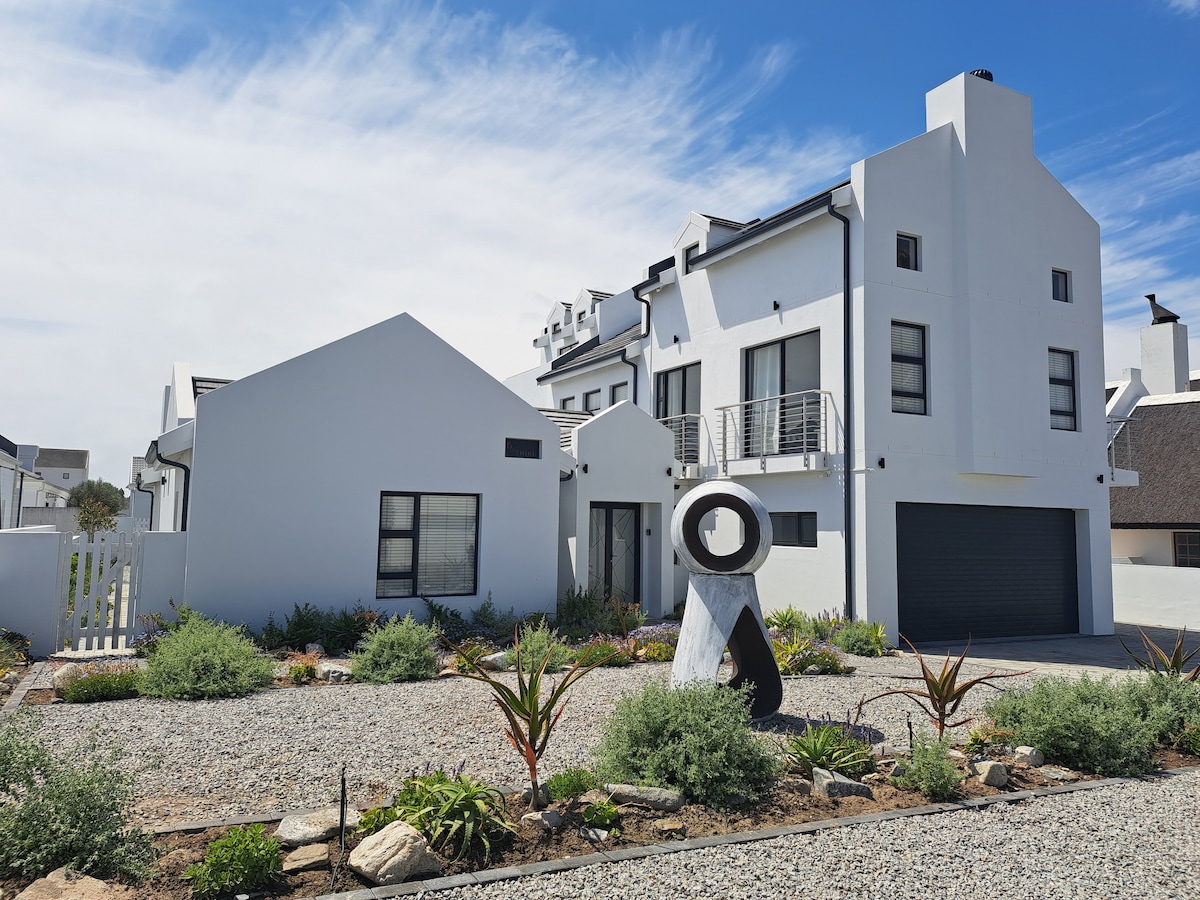 A modern, two-story home is set against a clear blue sky, featuring clean lines and a combination of textures. A sculptural piece is situated in a stone pathway, surrounded by low-maintenance greenery and decorative plants that enhance the landscape.