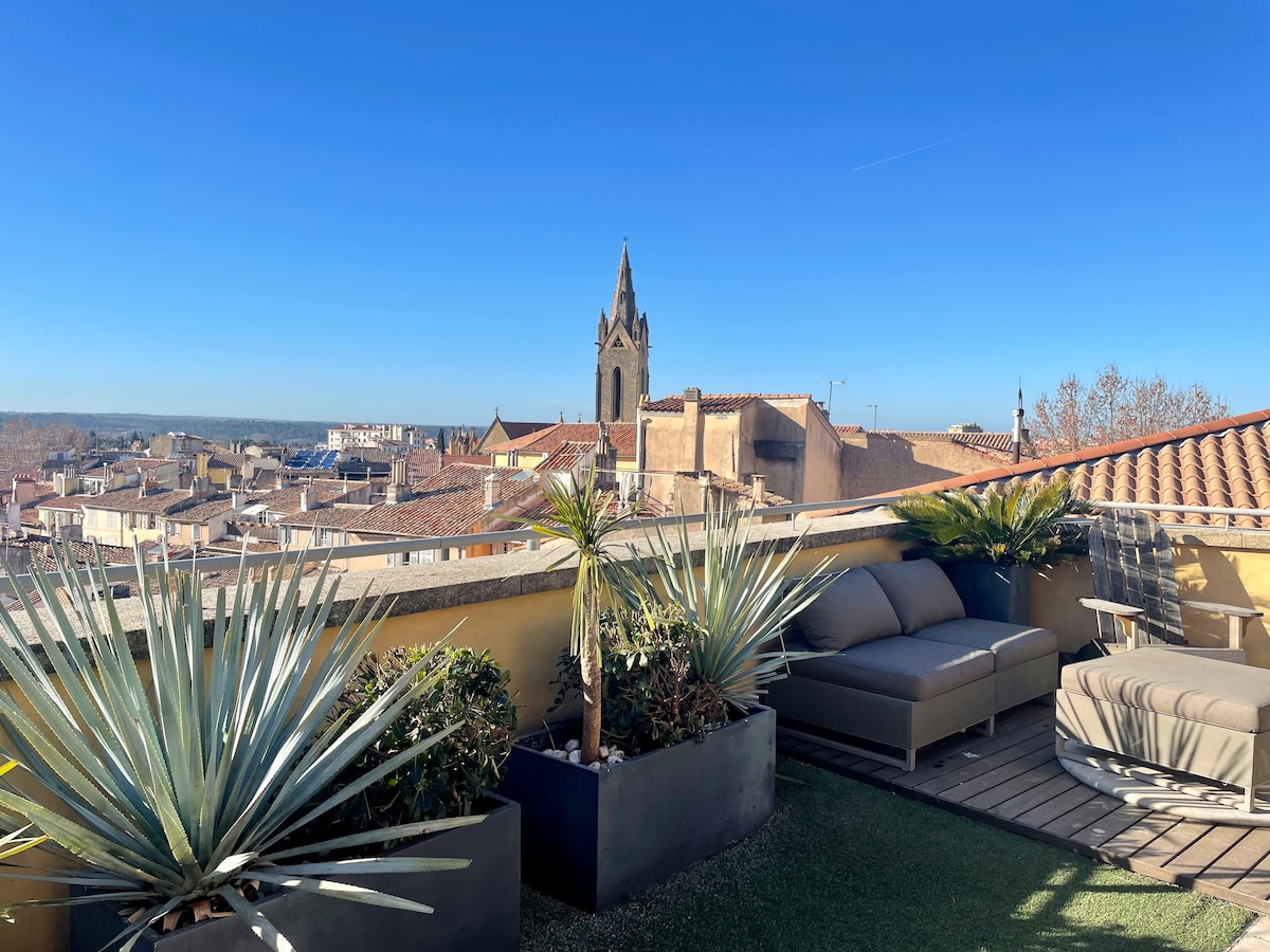A spacious terrace is displayed, featuring comfortable seating arrangements and potted plants. The view reveals rooftops of the city and a distant church steeple under a clear blue sky, providing a serene outdoor space for relaxation.