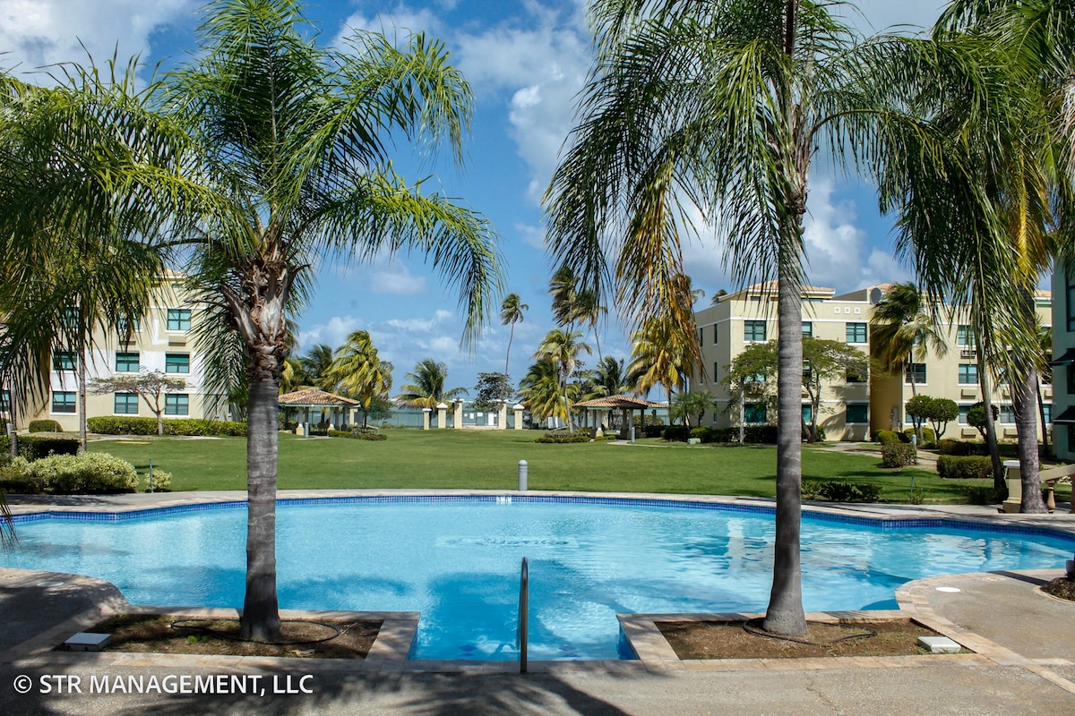A clear blue pool is framed by palm trees, with lush green lawns and shaded cabanas visible in the background. A serene tropical atmosphere is created by the bright sky and gentle landscape.