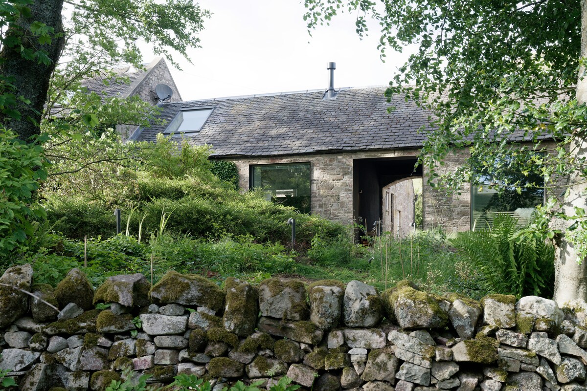 The exterior of a country cabin is framed by lush greenery, showcasing a blend of natural stone and wood elements. Large windows reflect the surrounding landscape, and a welcoming wooden door complements the rustic charm, with a neatly arranged stone wall visible in the foreground.
