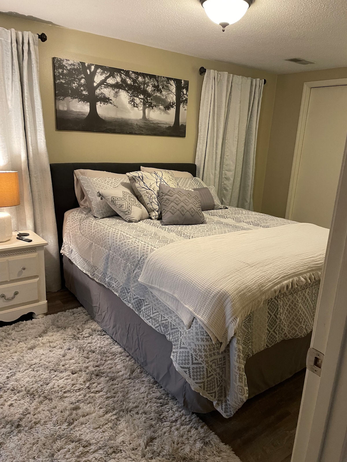 A cozy sleeping area is furnished with a spacious bed layered with light bedding and decorative pillows. A bedside table with a lamp is positioned next to the bed. Curtains frame the window, and a soft rug covers the floor.