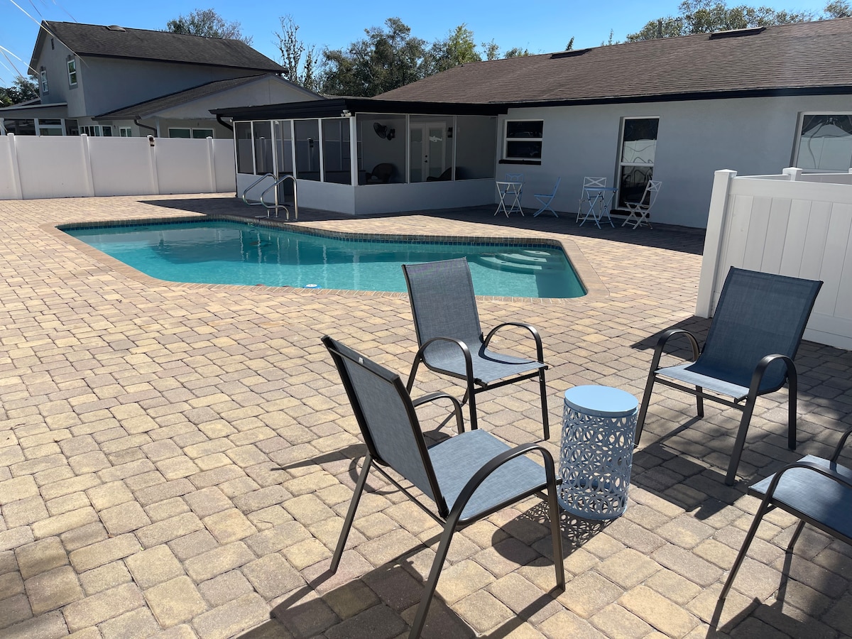 An outdoor space features a turquoise pool surrounded by a paved patio. Several gray chairs are positioned around a small table, offering a resting area. In the background, a covered seating area is visible alongside the house.