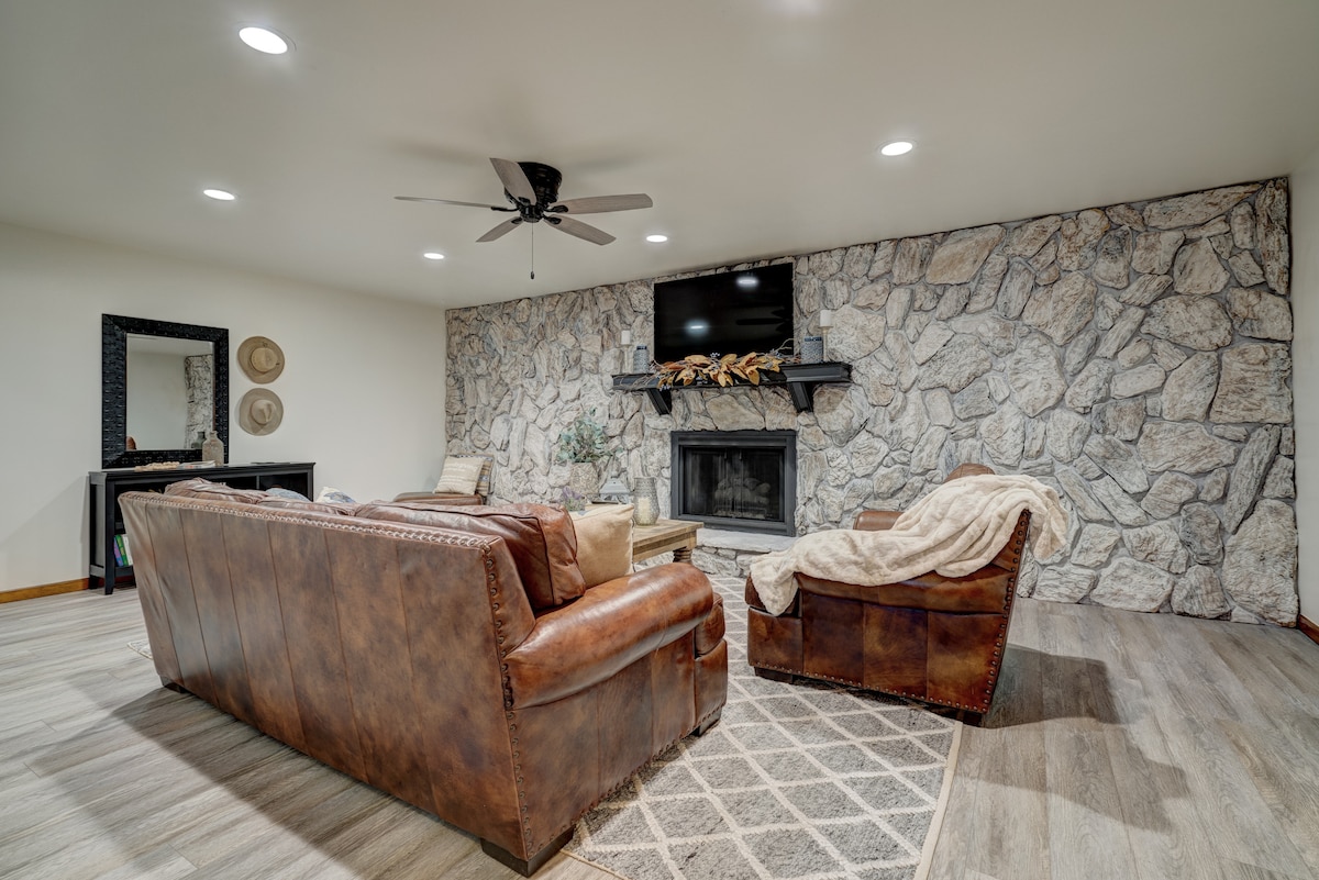 A cozy living area features a stone-accent wall, equipped with a mounted television above a fireplace. Two comfortable leather sofas are positioned around a textured area rug, complemented by a coffee table. Soft lighting creates a welcoming ambiance.