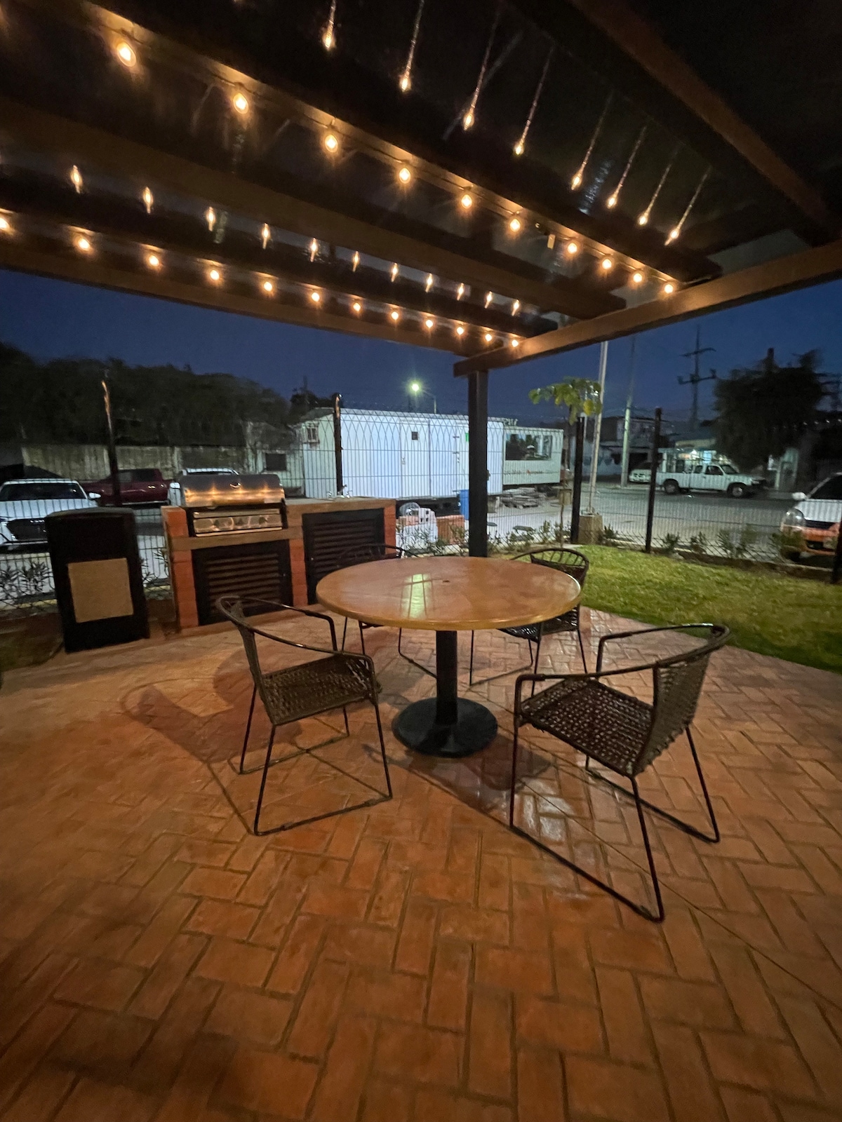 An outdoor seating area features a round table surrounded by two woven chairs. Soft lighting from overhead string lights creates a warm ambiance. In the background, a grill is visible, along with paved flooring and a view of the surrounding neighborhood.