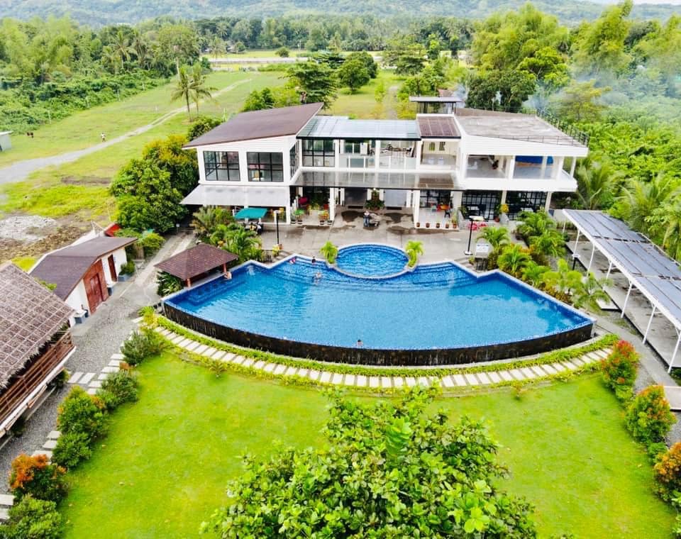 An aerial view showcases a large outdoor pool surrounded by a manicured lawn and lush greenery. The modern hotel structure features a mix of open and covered areas, with the inviting blue water of the pool contrasting with the vibrant landscape.