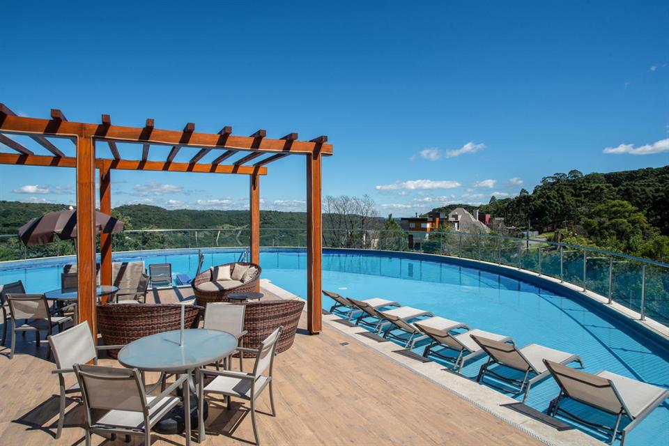 An outdoor terrace features a large swimming pool surrounded by lounge chairs, providing scenic views of the surrounding landscape. A shaded seating area with a circular wicker couch and tables is positioned under a wooden pergola, inviting relaxation.