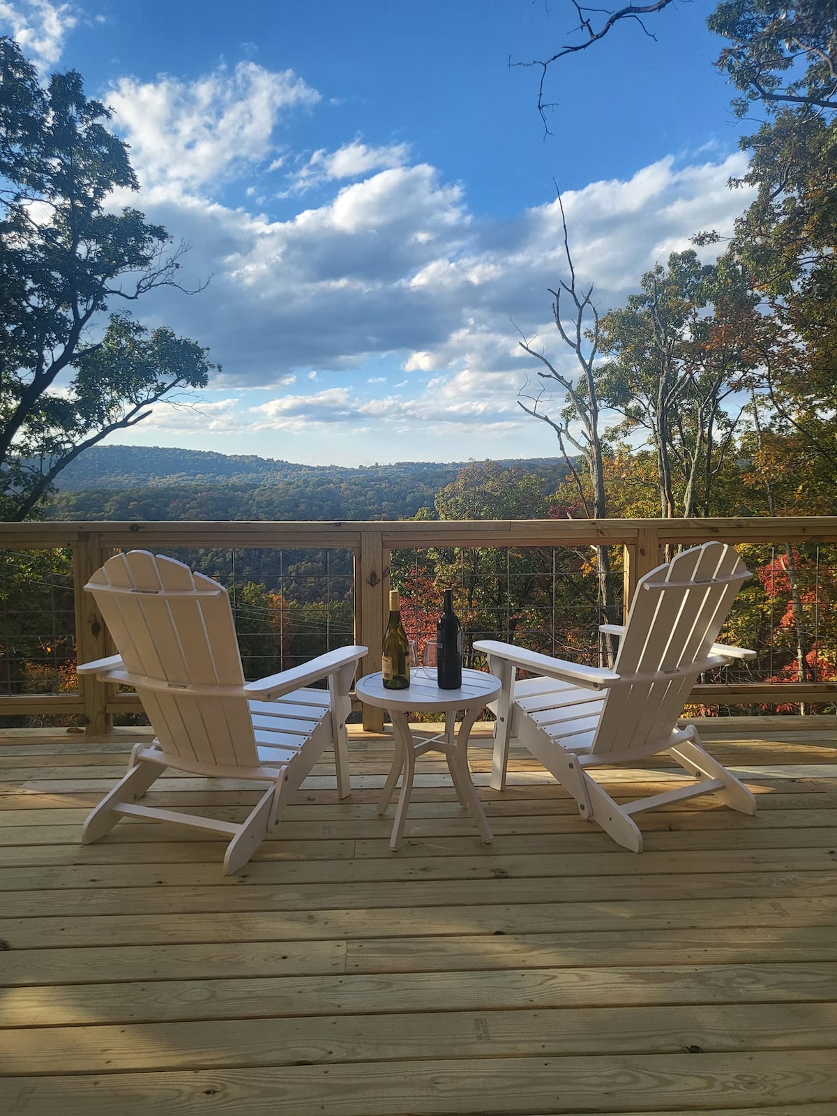 Two white Adirondack chairs and a small side table are positioned on a wooden deck. A scenic view of rolling hills and trees with autumn foliage is visible in the background, under a partly cloudy sky.