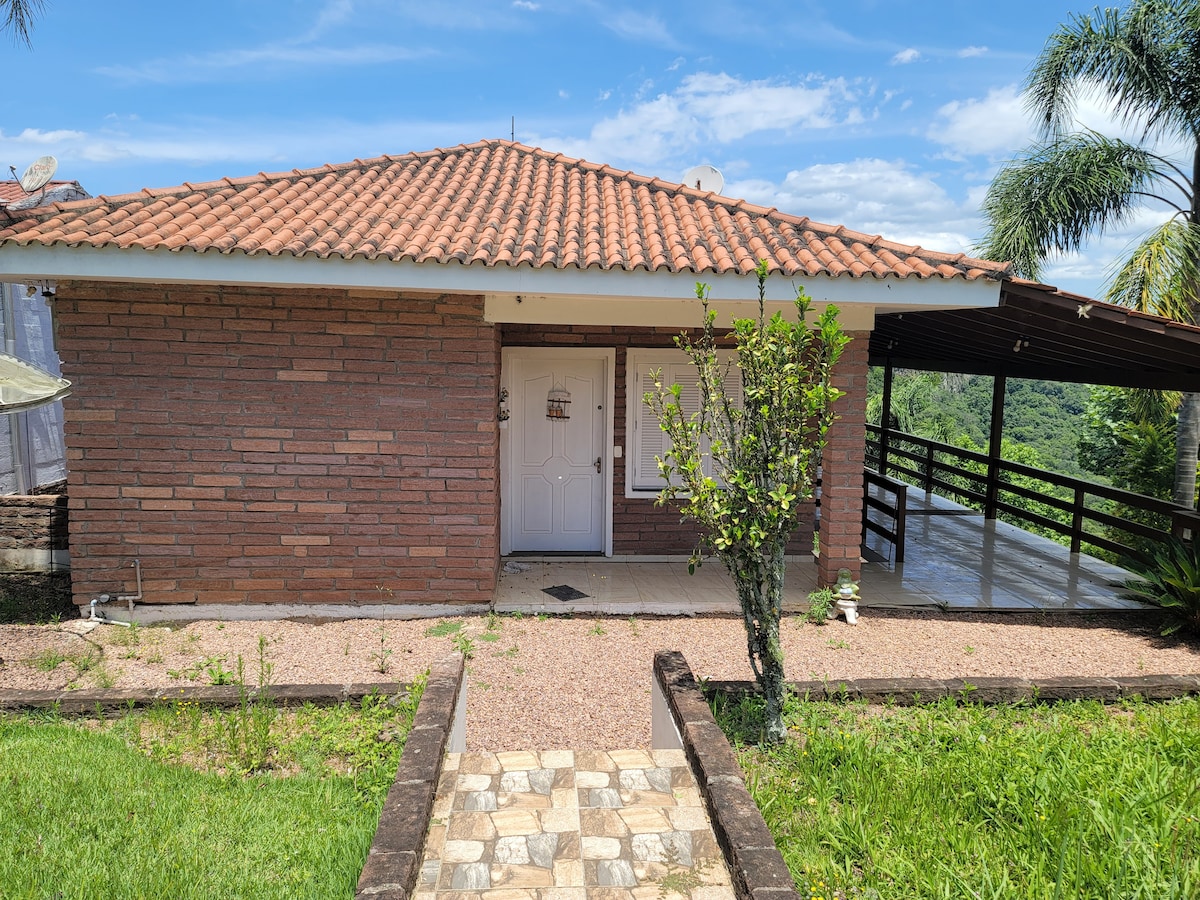 A single-story brick house is shown, featuring a tiled roof and a white front door. A pathway of stones leads to the entrance, surrounded by grassy areas and a small shrub. A covered porch extends alongside the house, with a view of trees in the background.