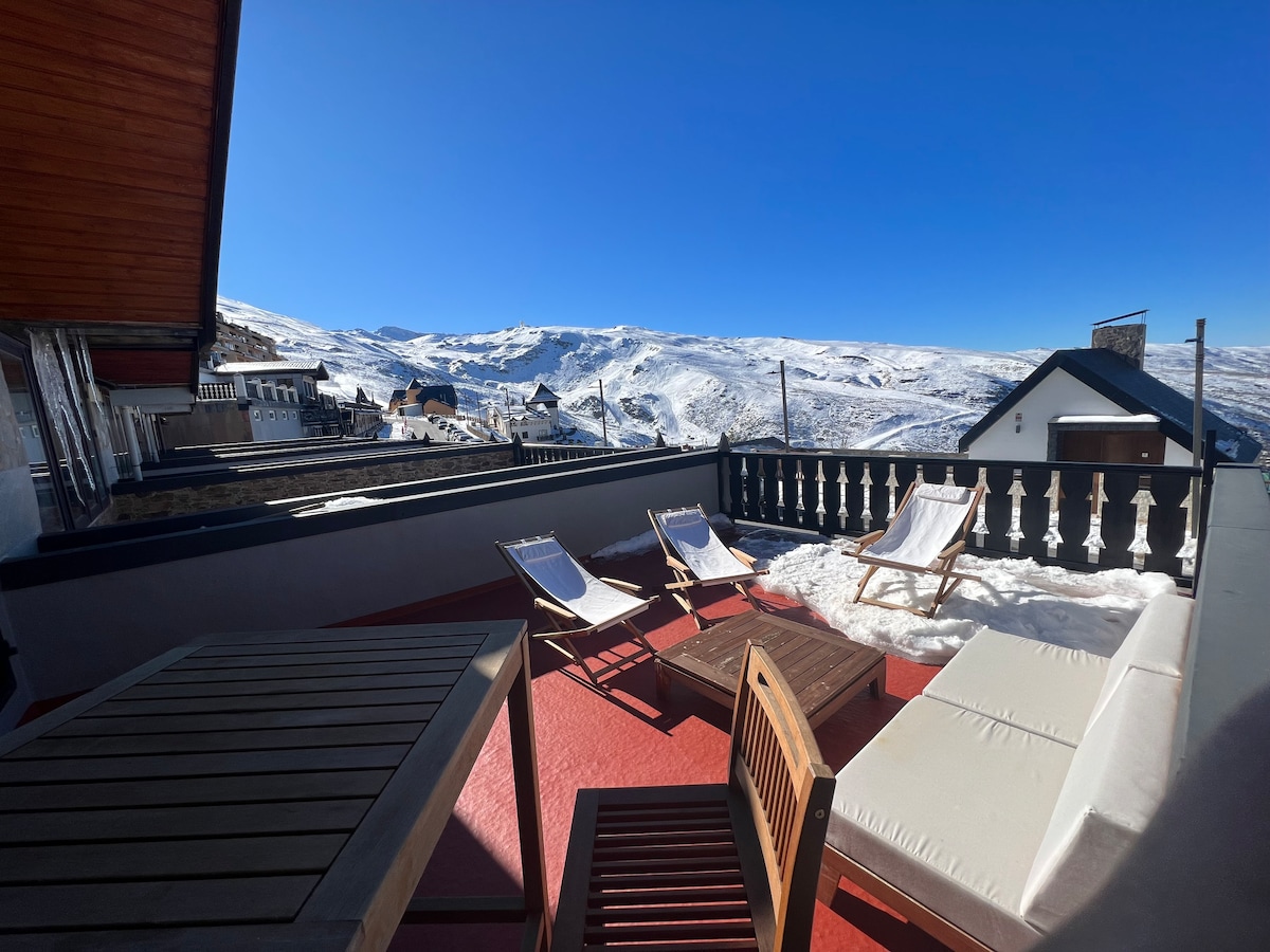 A spacious terrace is shown, featuring sun loungers and a wooden table. The panoramic view includes snow-covered mountains and a clear blue sky. The terrace is highlighted by red flooring, suggesting a comfortable outdoor space for relaxation.