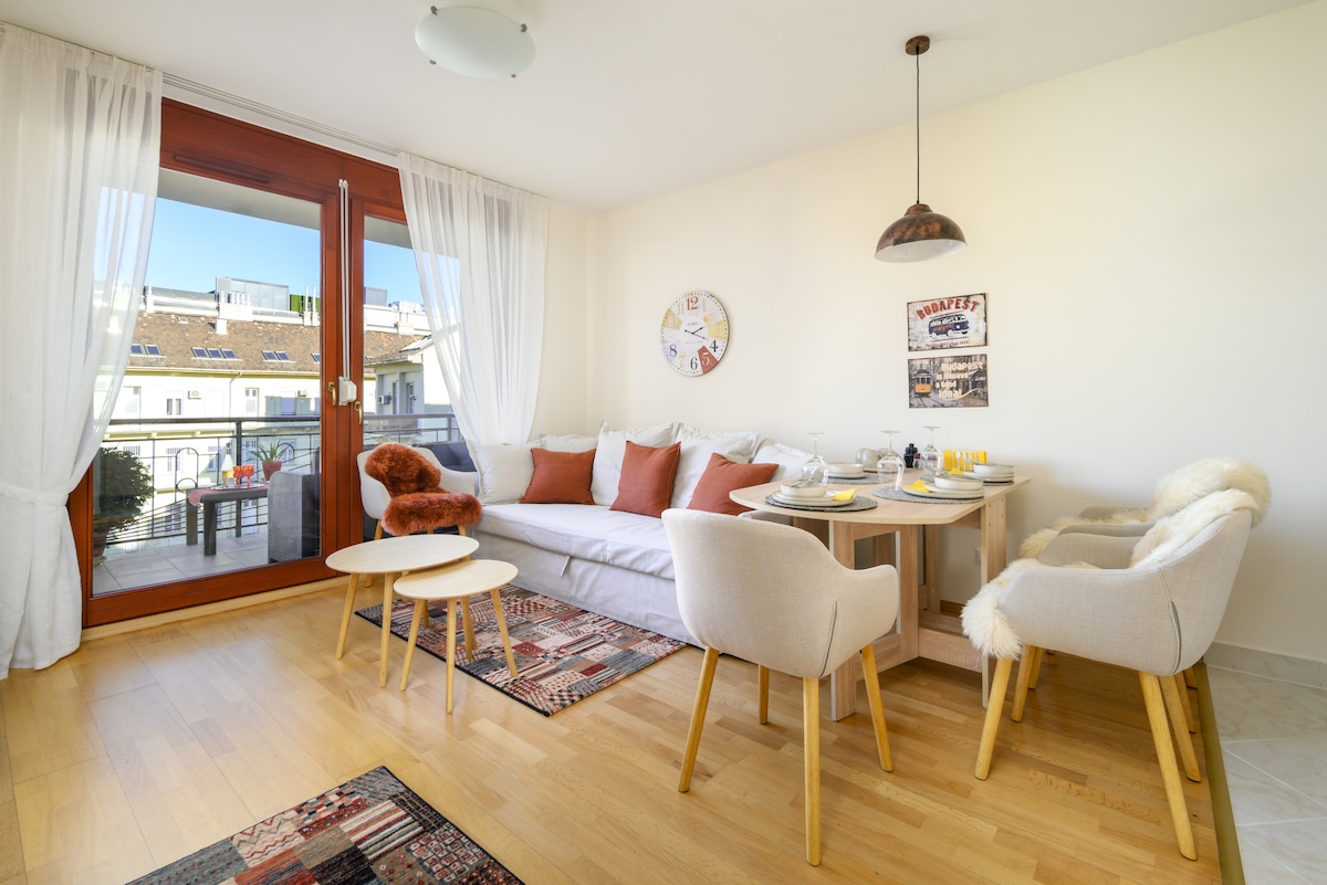 A bright living area features a cozy sectional sofa adorned with plush cushions, accompanied by two small tables. A dining space is visible with a set of four chairs and a tabletop neatly arranged for a meal. Large glass doors open to a balcony with outdoor seating.
