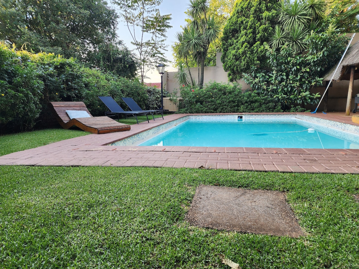 A tranquil outdoor space features a refreshing pool surrounded by manicured grass and lush greenery. Two lounge chairs are positioned beside the pool, inviting relaxation. The area is complemented by well-maintained plant life, creating a calming atmosphere.