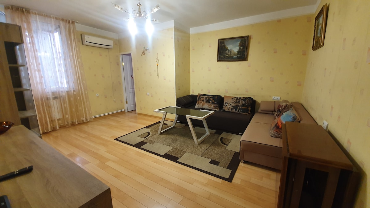 A spacious living area showcases a large sectional sofa accompanied by a contemporary coffee table. Soft lighting is provided by overhead fixtures and natural light filters through sheer curtains. A decorative rug adds warmth to the wooden floor, while artwork is displayed on the wall.