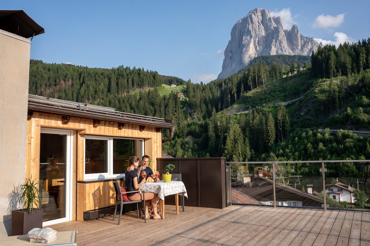 A private terrace features a wooden table set with two chairs, providing a serene outdoor dining experience. In the background, a striking mountain landscape is visible, complemented by lush greenery and a clear blue sky.