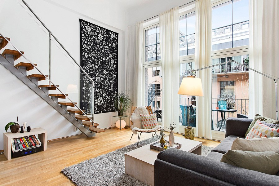 A spacious living area features a soft carpet and a comfortable sofa adorned with decorative pillows. Large windows provide ample natural light and views of the balcony, while a stylish staircase leads to the lofted space above.