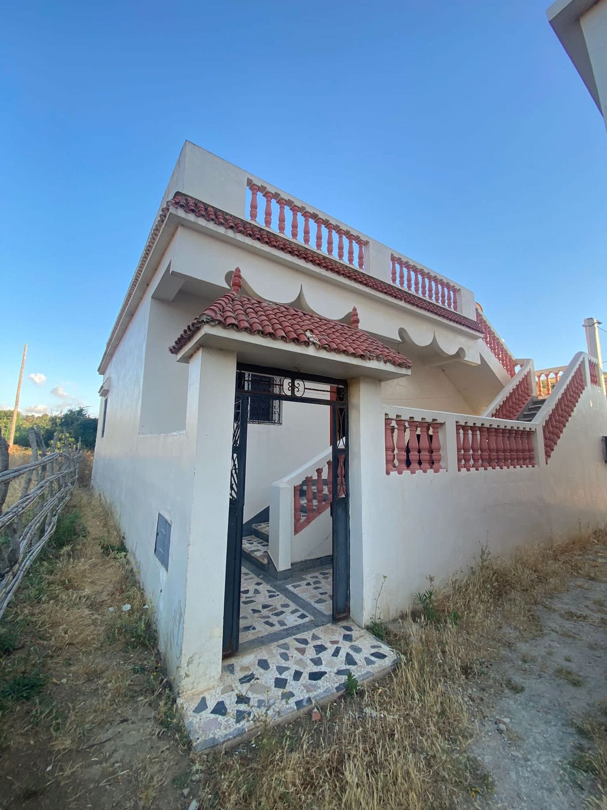 A small villa is depicted, featuring a distinctive architectural style with a tiled roof and decorative elements. An entrance gate opens to a pathway lined with stone tiles. A staircase leads to an upper level, while surrounding grassy areas contribute to the serene environment.