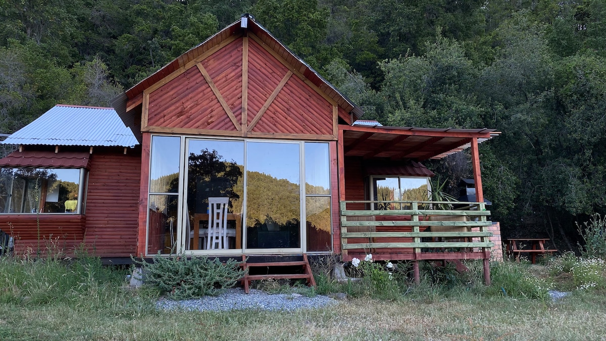 A rustic cabin features a bright red exterior and large glass windows allowing views of the surrounding landscape. A porch with a railing and a seating area is visible, alongside a grassy yard with patches of greenery and gravel.