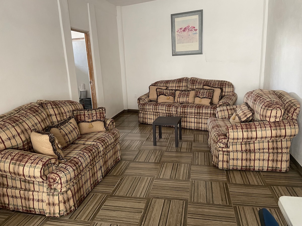 A spacious living area features multiple patterned sofas arranged around a small table. Natural light fills the space through a nearby doorway, while the patterned floor tiles add visual interest to the room.