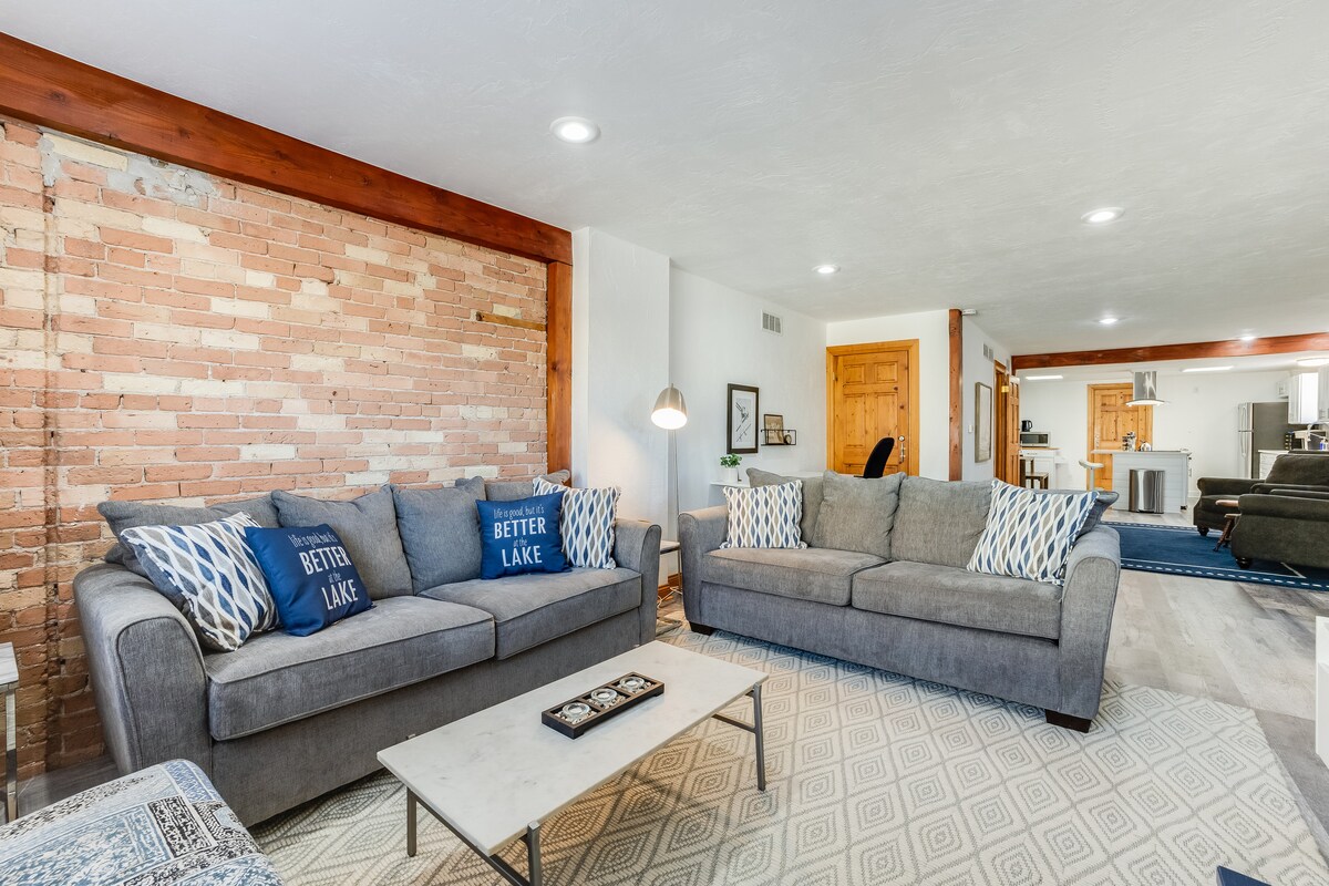 A spacious living area features comfortable gray sofas with decorative cushions, a light wooden coffee table, and an area rug underfoot. Exposed brick walls add texture, while soft lighting enhances the warm atmosphere. A glimpse of the dining area is visible in the background.