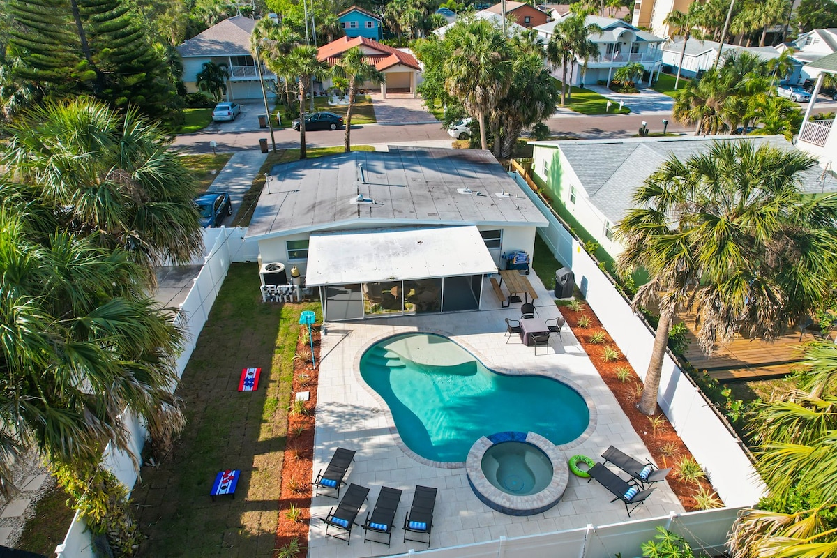 An aerial view of a beach cottage property showcases a heated saltwater pool surrounded by lounge chairs. A spa section is integrated into the pool design. The backyard is enclosed with a privacy fence, featuring landscaped areas and outdoor seating options.