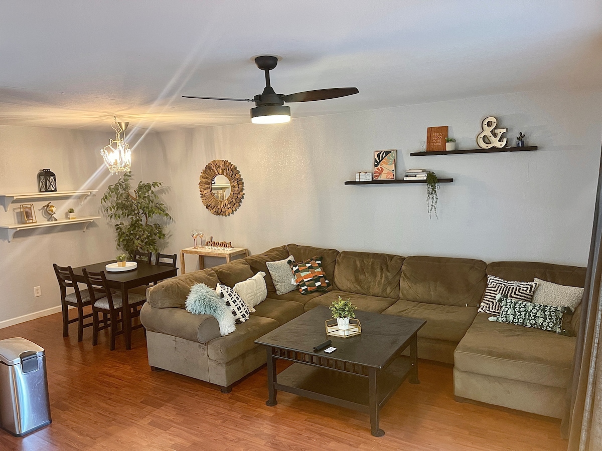 A comfortable living area is furnished with a large sectional sofa and a coffee table. A dining table with chairs is positioned nearby, while a decorative shelf displays various items. Natural light is provided by an overhead fan and fixtures, enhancing the cozy ambiance.